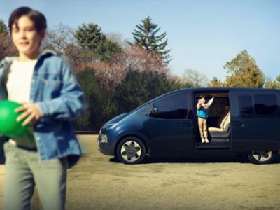 Ein Junge mit Ball. Seine Schwester winkt ihm aus einem Hyundai STARIA heraus nach. 