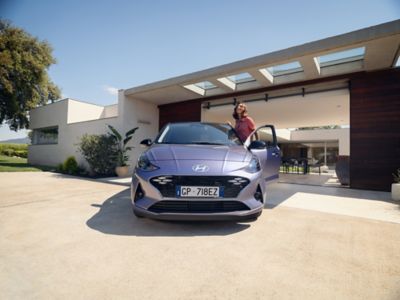 Ragazza sorride sulla porta di Hyundai i10 davanti a una casa lussuosa e moderna