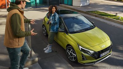 Person standing beside a Hyundai i20 parked near modern buildings.