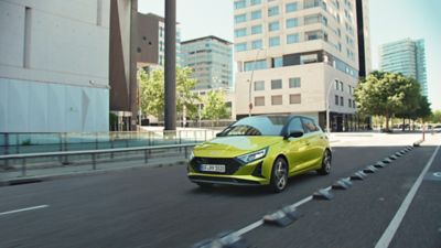 A green Hyundai i20 driving between modern building blocks.	