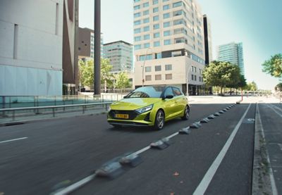 A green Hyundai i20 driving between modern building blocks.	