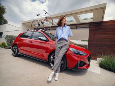 A woman leaning on a red i30 Hatchback with a Hyundai Genuine Accessories roof top bike carrier.