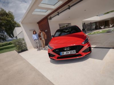 A man an woman walking next to the Hyundai i30 N Line parked in front of a house.