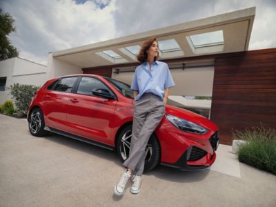 Une femme se tient devant une Hyundai i30 N Line rouge, garée devant une maison.