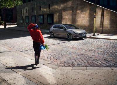 Vrouw in rode trui loopt naar geparkeerde Hyundai i30 driekwart van voren.