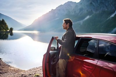 Man die tegen zijn Hyundai i30 N Line Wagon aanleunt en naar een meer kijkt.  looking out on a mountain lake