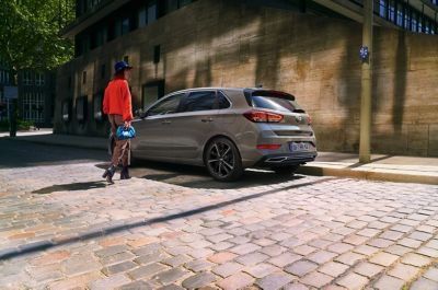 Eine Frau in roter Jacke geht zu ihrem am Straßenrand geparkten grauen Hyundai i30.