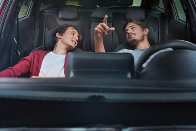 Padre e hijo sentados en la parte delantera de un nuevo Hyundai i30 Fastback aparcado. Sus asientos están reclinados y están hablando entre ellos.
