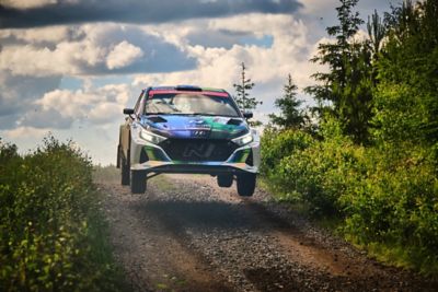 A Hyundai N racing car jumps on a gravel track, surrounded by trees and a cloudy sky.