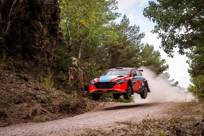 The Hyundai i20 N WRC Rally2 challenger drifting around a corner on a dirt road.