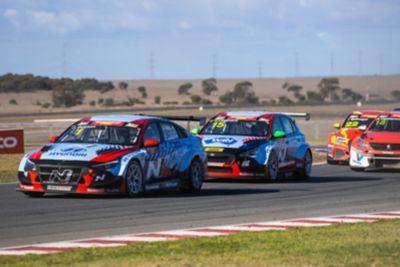 Hyundai N race cars speed on a track, with a scenic landscape and powerlines in the background.