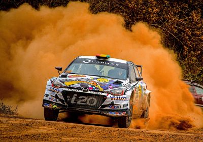 A Hyundai i20 N racing car powers through a dirt track, kicking up a massive cloud of dust.