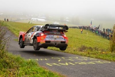 The Hyundai i20 N racing car jumps during a rally, with spectators and foggy fields around.