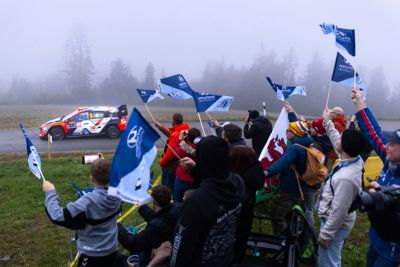 A Hyundai N racing car speeds along a foggy rally track as fans cheer and wave flags enthusiastically.