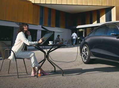 A lady charging a laptop via the exterior charging port of the Vehicle-to-Load technology of IONIQ 6.