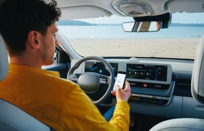 Ein Mann am Steuer eines am Strand parkenden Hyundai Fahrzeugs mit einem Smartphone in der rechten Hand.