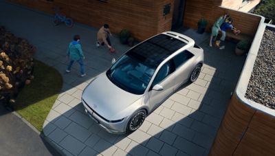 Silvrig Hyundai IONIQ 5 parkerad på uppfart i bostadsområde, sedd uppifrån med personer i bakgrunden.