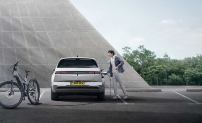 The Hyundai IONIQ 5 electric vehicle pictured from in front modern building with a man using V2L to charge electric bike.