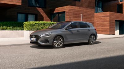 Vista a tre quarti dalla parte frontale di nuova Hyundai i30 Hatchback di colore grigio, parcheggiata di fronte a un’abitazione in stile moderno