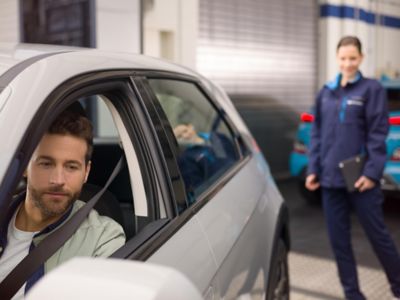 A man driving a Hyundai i30 away from a Hyundai dealership with a woman in the background.	