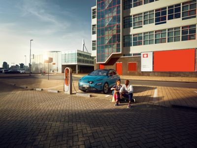Una coppia seduta in un parcheggio mentre Nuova Hyundai Kona Electric è in carica presso una stazione di ricarica pubblica. 