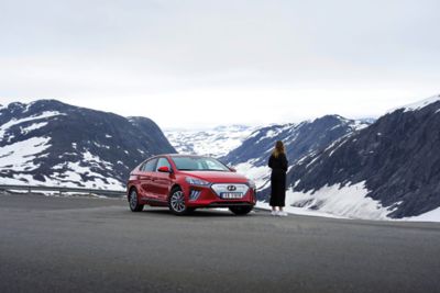En dame står foran en Hyundai IONIQ Electric parkert i et fjellandskap. Foto.