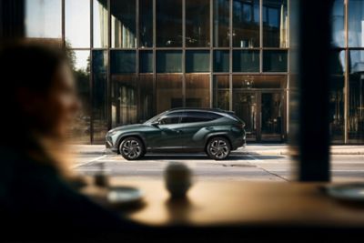Hyundai TUCSON parkerad utanför modern byggnad med glasfasad, sedd genom ett fönster.