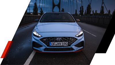 Der Hyundai i30 N in der Dämmerung auf einer Brücke mit eingeschalteten Scheinwerfern.