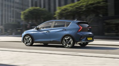 Side view of a blue Hyundai BAYON driving down a city street with trees in the background.