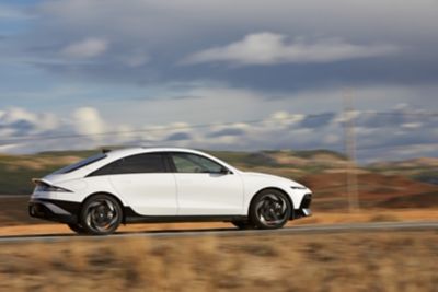 Zijaanzicht Hyundai IONIQ 6 rijdend op een weg in een heuvelachtige omgeving.