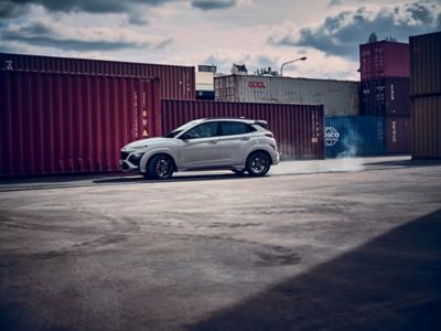 Vue latérale du SUV sportif Hyundai KONA N évoluant à toute vitesse entre les conteneurs d’un site industriel.