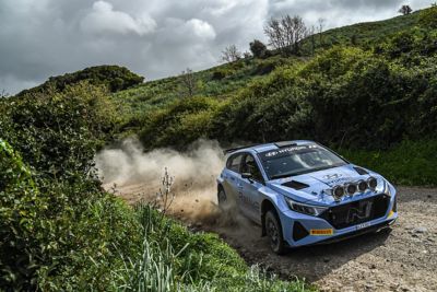 Ein Hyundai i20 N Rally2 fährt auf einer staubigen Rennstrecke umgeben von Wiese und Gebüsch.