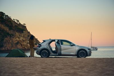 A beach scenery and the IONIQ 5 about to charge. A woman is standing next to the car.