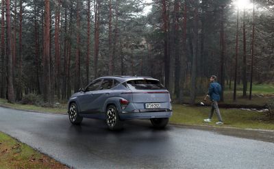 Nowy model Hyundai KONA stoi zaparkowany na leśnej drodze, a obok idzie mężczyzna.