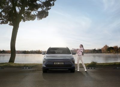 The all-new Hyundai KONA electric parked in front of a lake with a woman standing next to it.