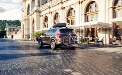 Hyundai SANTA FE Hybrid z zamontowanymi belkami dachowymi, boksem dachowym oraz bagażnikiem rowerowym na hak holowniczy.