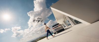 Hyundai IONIQ 5 à côté d’un bâtiment moderne, un véhicule de mobilité aérienne urbaine volant dans le ciel.