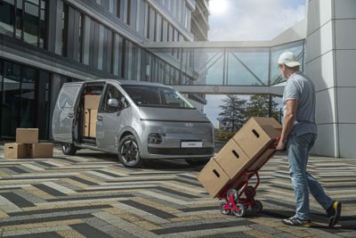 Driekwart vooraanzicht Hyundai STARIA Cargo met geopende schuifdeur en man die met steekkar dozen naar de elektrische bestelwagen rijdt.