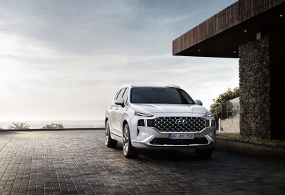 The Santa Fe 7 seat SUV in white from the front parked in front of a house with ocean view.