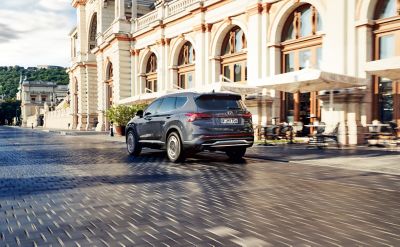 The new Hyundai Santa Fe Hybrid 7 seat SUV in grey driving down a city street.