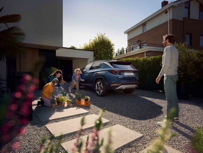 The Hyundai TUCSON Plug-in Hybrid parked next to its home charging box and a family playing.