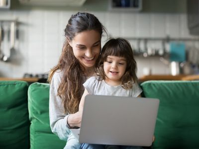 Matka z małą córeczką siedzą na zielonej kanapie w domu i korzystają z laptopa.