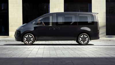 Der Hyundai STARIA Hybrid in der Seitenansicht, vor einem Bürogebäude parkend. 