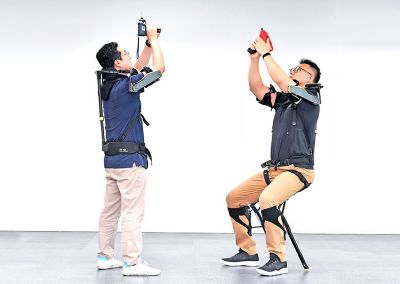 Dwóch mężczyzn wypróbowuje kamizelkę egzoszkieletową VEX firmy Hyundai w warunkach roboczych.