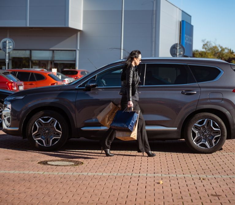 Žena kráčajúca s nákupom do svojho šedého Hyundai SANTA FE Hybrid zaparkovaným pred nákupným centrom