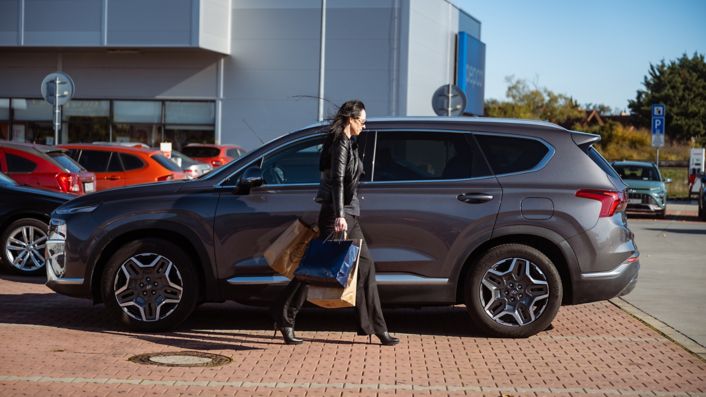 Žena kráčajúca s nákupom do svojho šedého Hyundai SANTA FE Hybrid zaparkovaným pred nákupným centrom