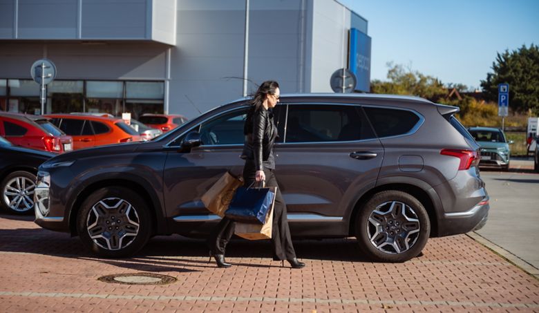 Žena kráčajúca s nákupom do svojho šedého Hyundai SANTA FE Hybrid zaparkovaným pred nákupným centrom