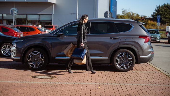 Žena kráčajúca s nákupom do svojho šedého Hyundai SANTA FE Hybrid zaparkovaným pred nákupným centrom