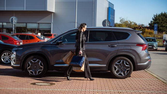 Žena kráčajúca s nákupom do svojho šedého Hyundai SANTA FE Hybrid zaparkovaným pred nákupným centrom