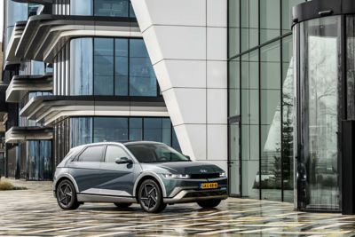 Driekwart vooraanzicht geparkeerde Hyundai IONIQ 5 voor een modern gebouw met veel glas.